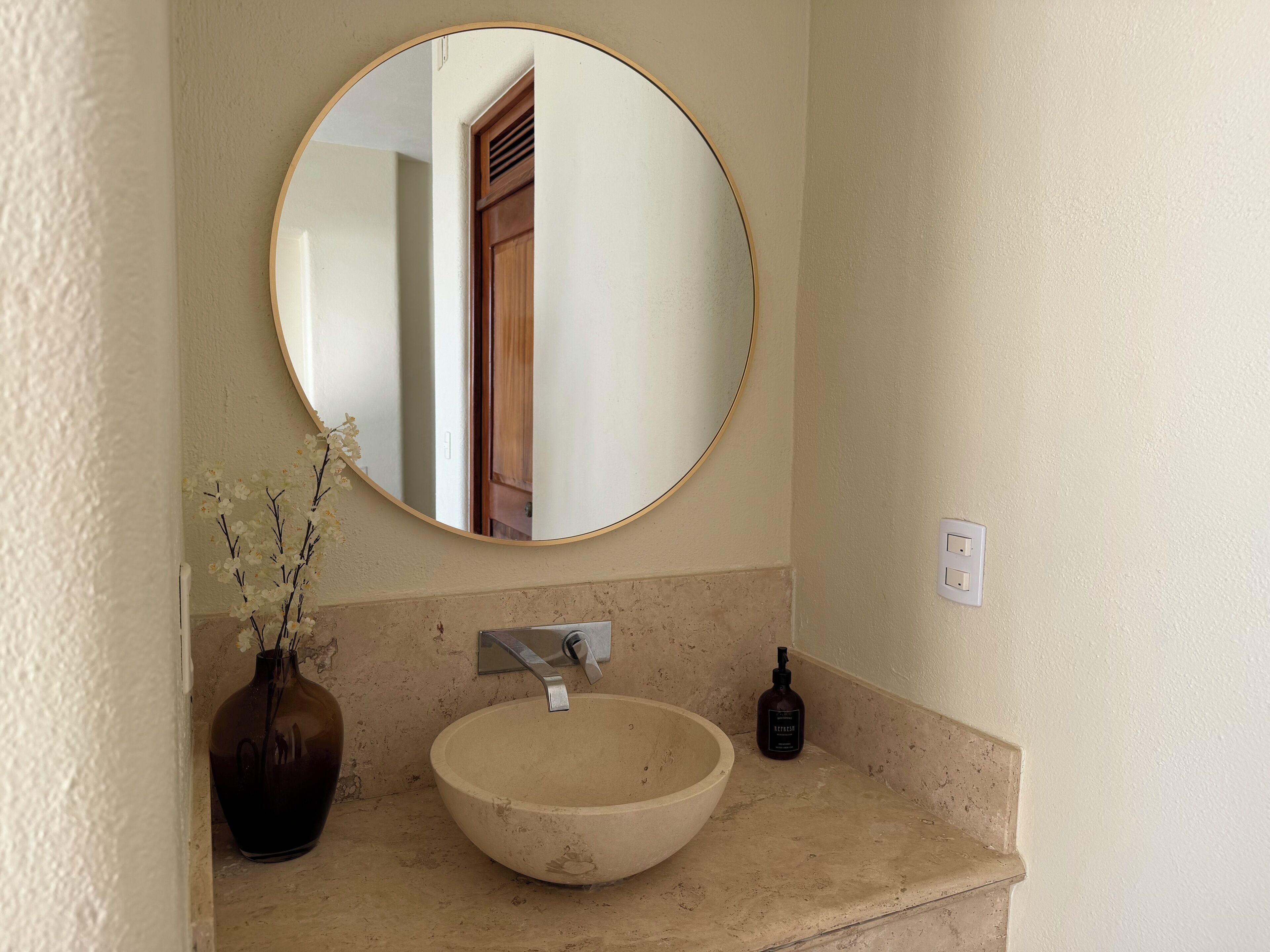 Un baño con un espejo redondo, un lavabo color beige, un florero oscuro con flores y una botella de jabón líquido.