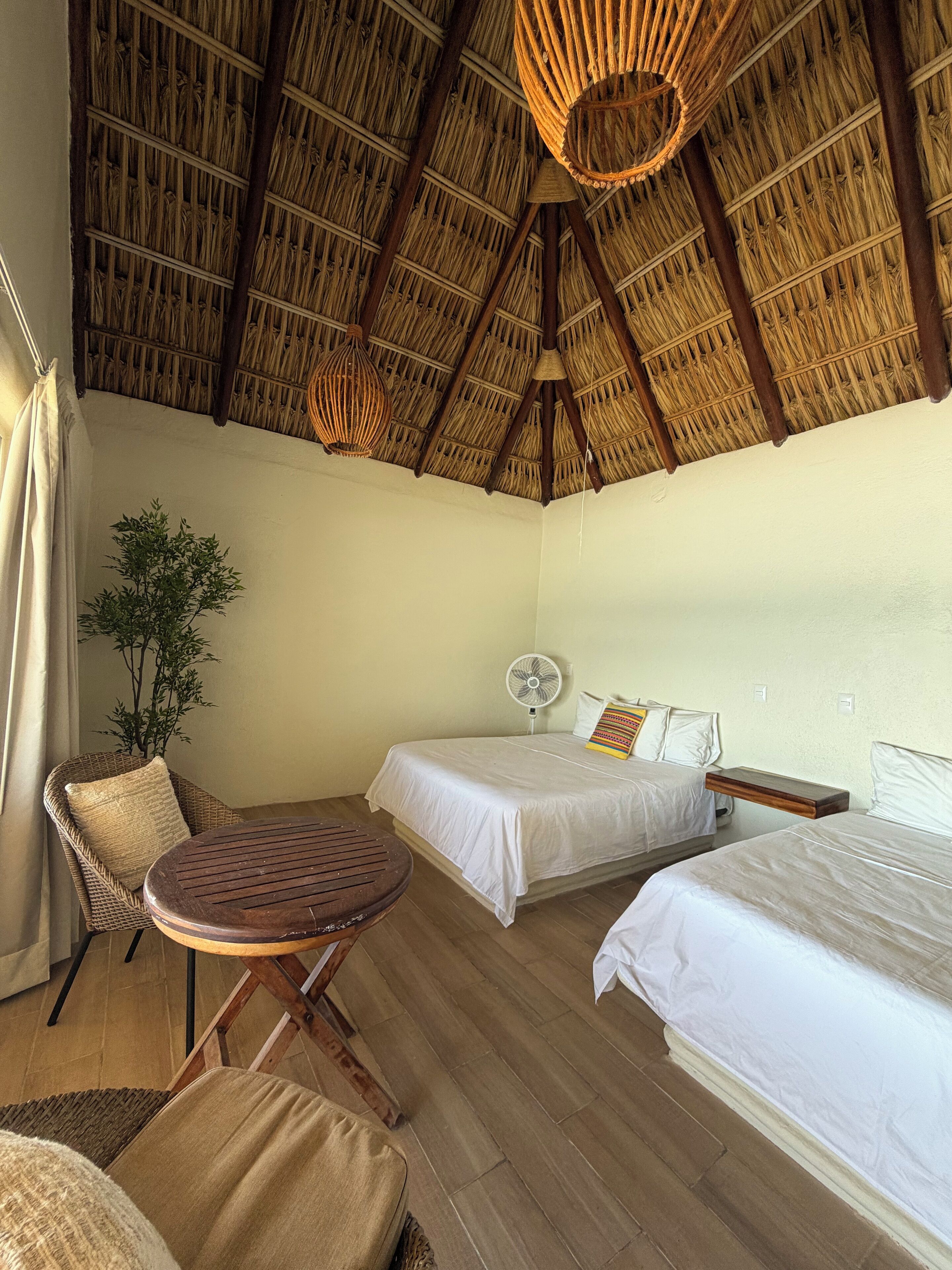 A room with two beds, a fan, a wicker chair, a small table, and a ceiling with a thatched roof.