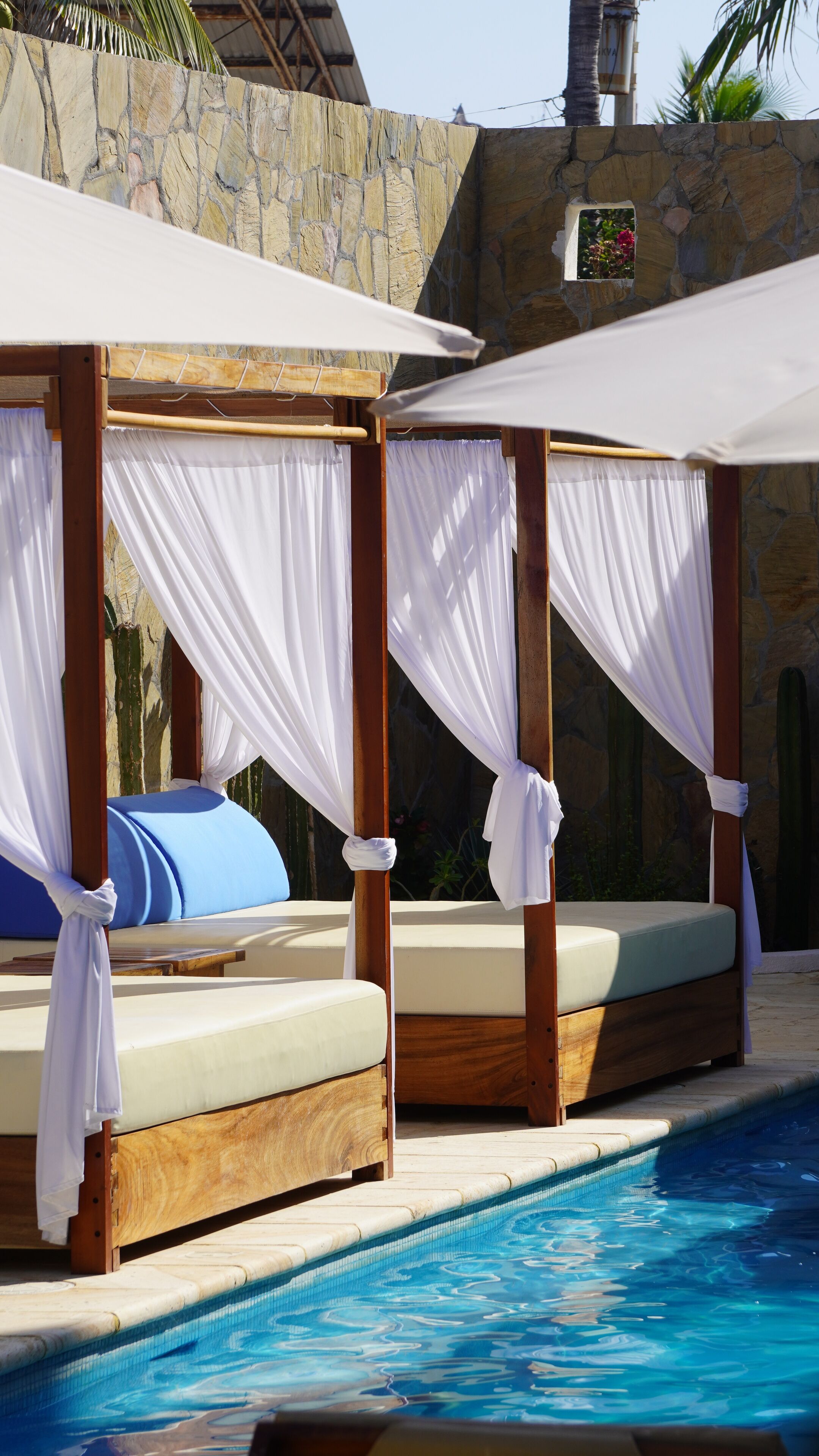 Dos cabañas de madera con toldos blancos y cojines junto a una piscina.