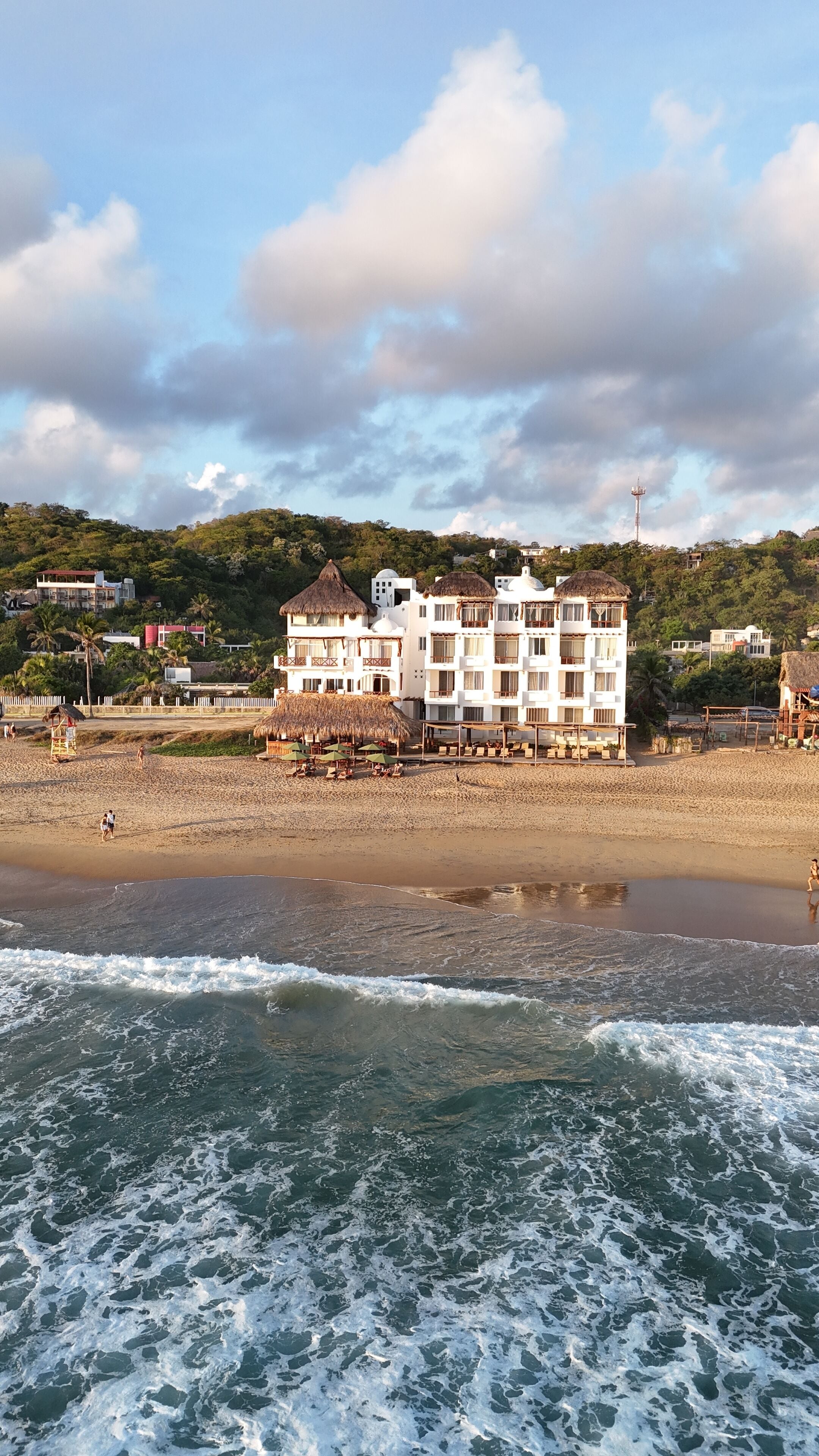 A beachfront hotel with multiple floors, a beach in the foreground, and a hill with greenery and buildings in the background.