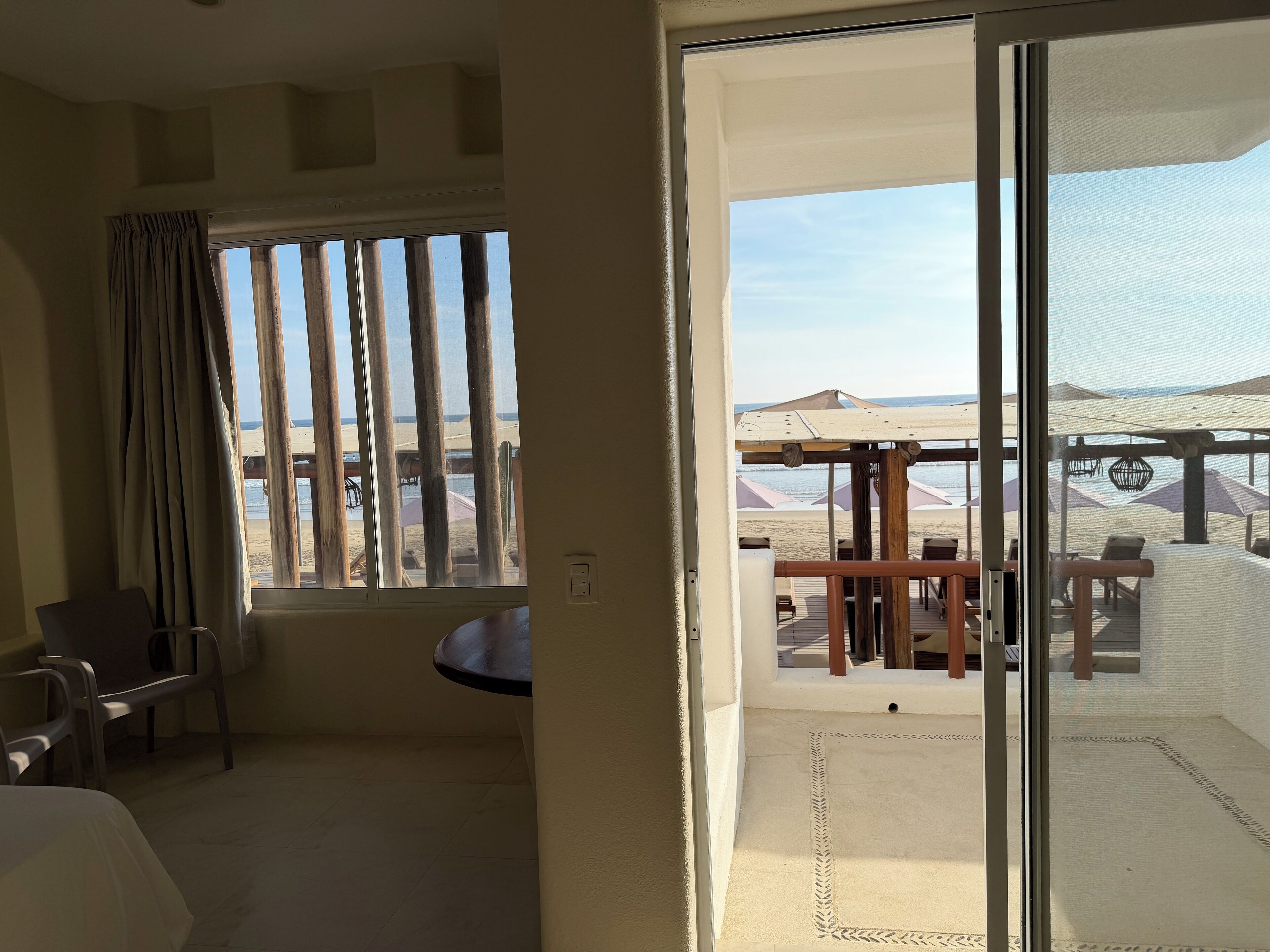 A room with a balcony overlooking a beach, featuring a table and chairs, a view of the beach, and a large window with curtains.