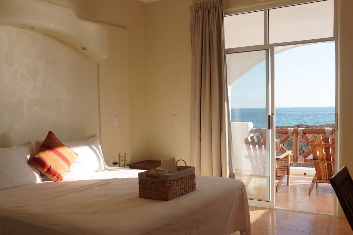 Habitación de hotel con cama, mesita de noche, cesta, silla y vista al mar a través de un ventanal.