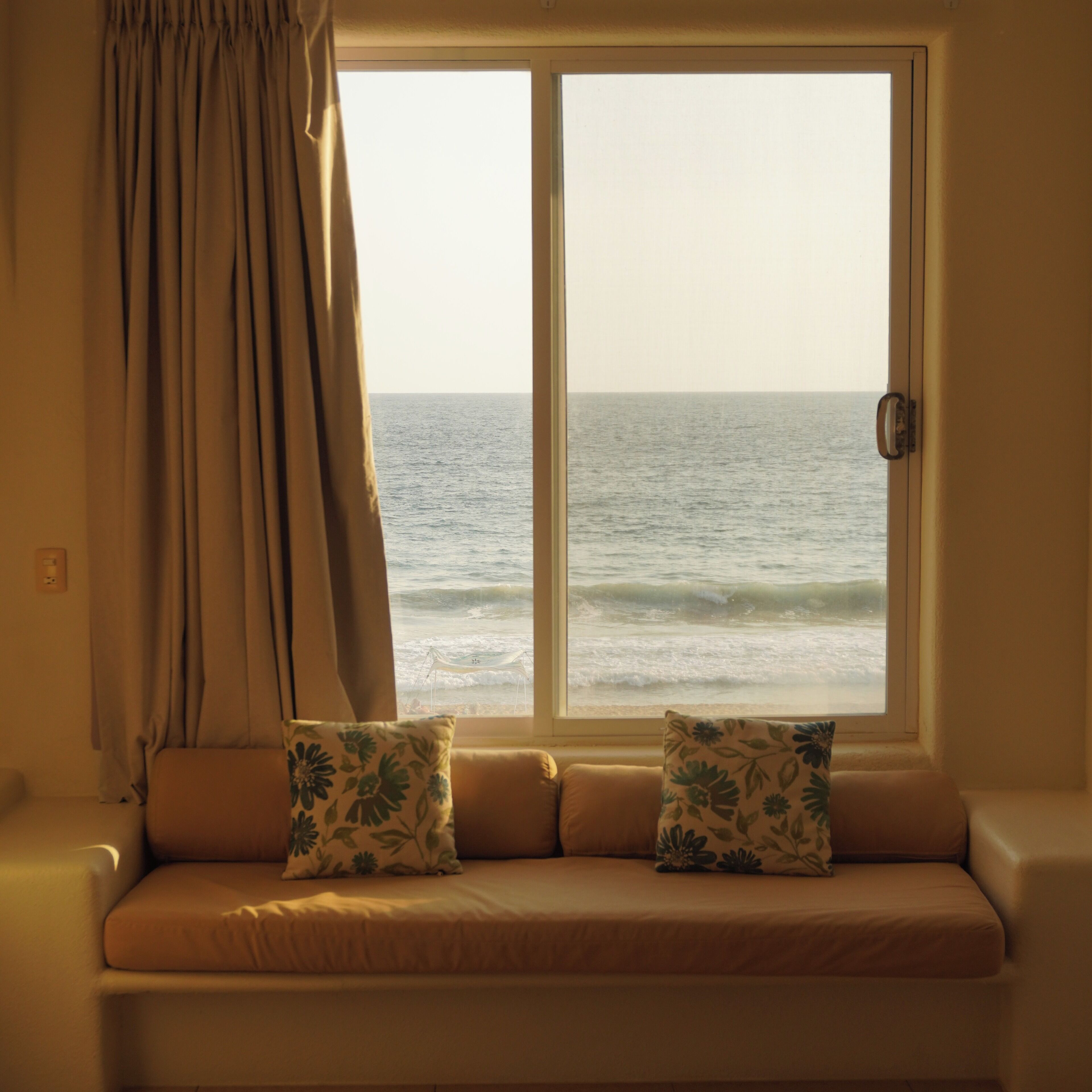 A window with a view of the ocean, a sofa with floral cushions, and a curtain.
