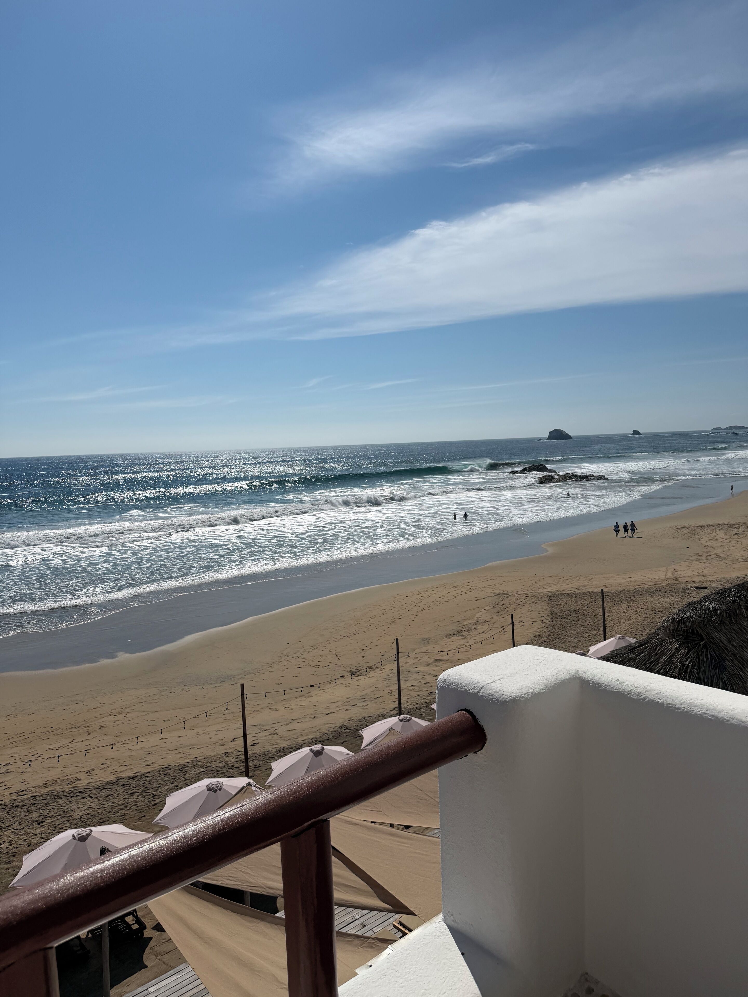 A beach view from a balcony with a railing, beach umbrellas, and a sandy shore.