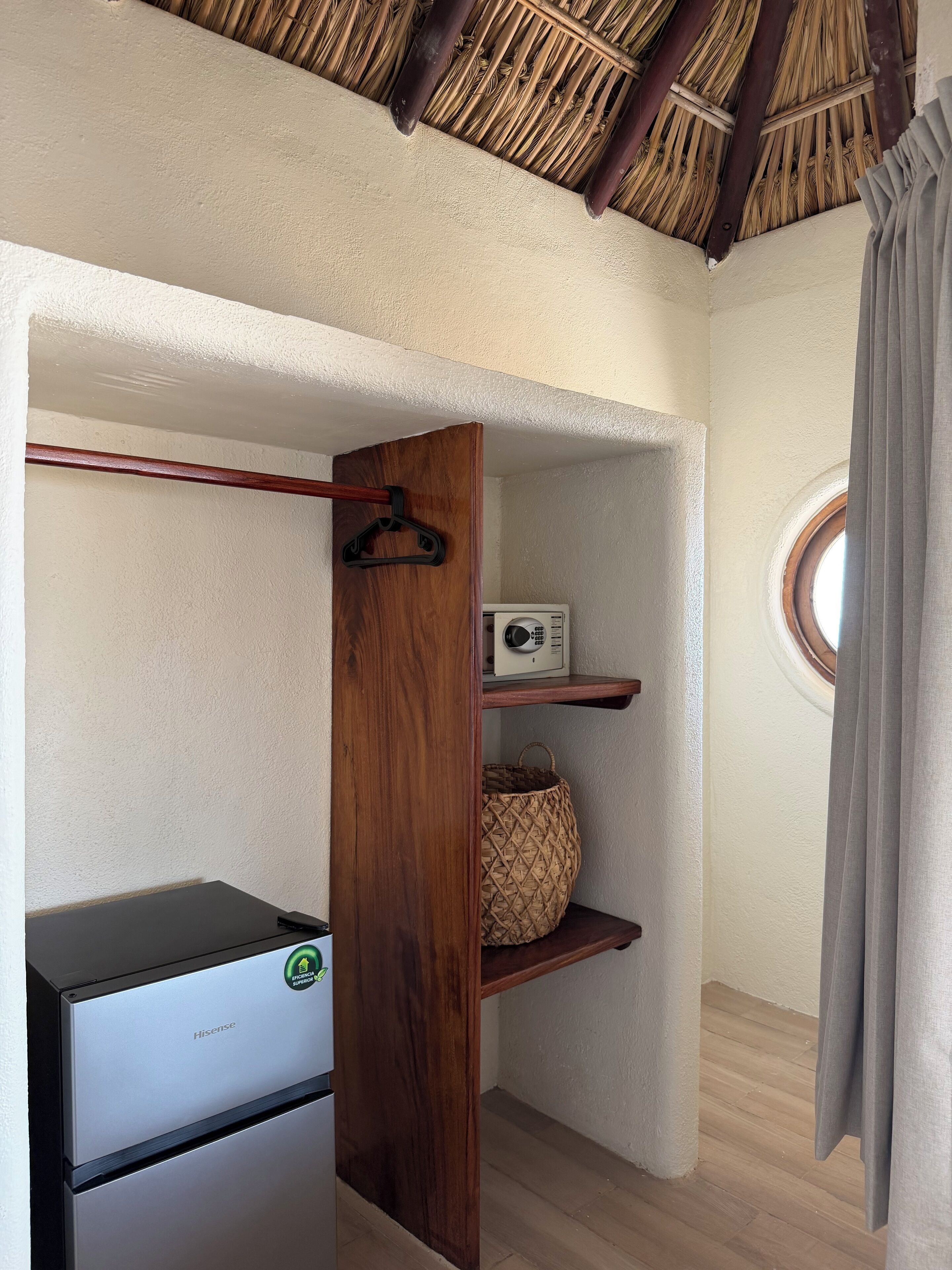 A compact room with a wooden wardrobe, a small shelf with a woven basket, a white appliance, and a window with a view.