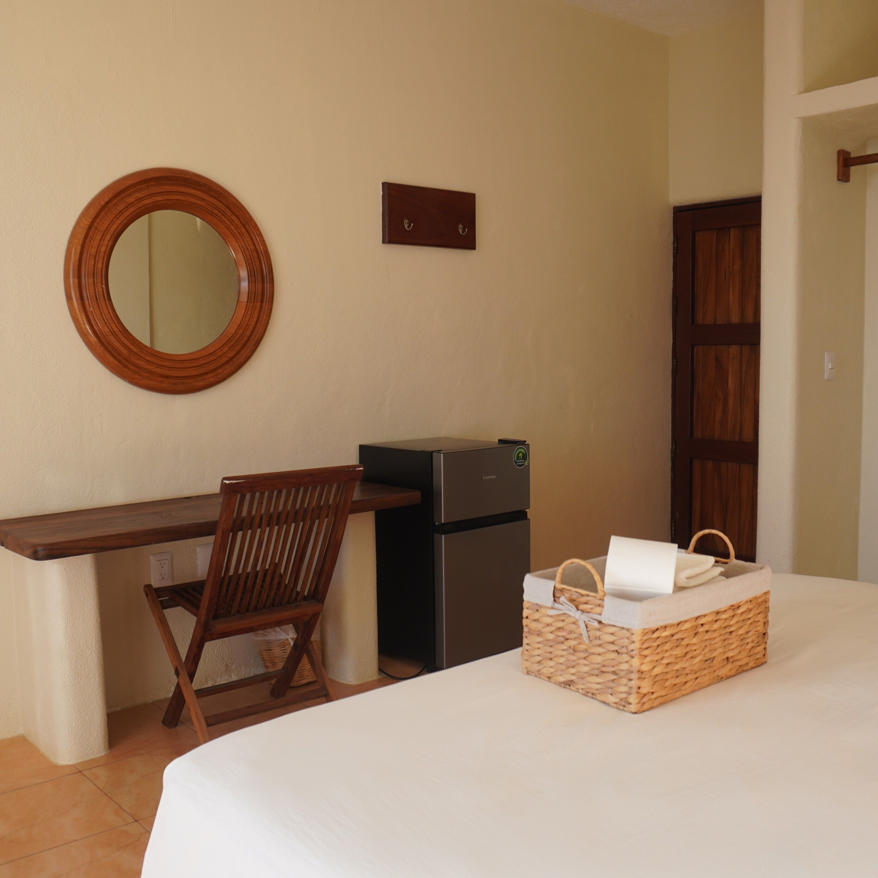 Une chambre d’hôtel avec un lit, un bureau et une chaise, un petit réfrigérateur, un panier en osier et une porte en bois.