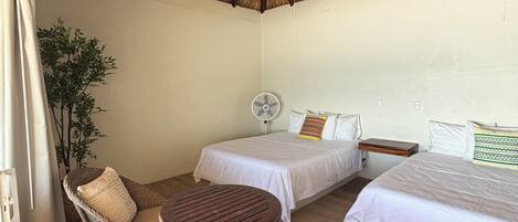 Una habitación con techo de paja, dos camas, un ventilador, mobiliario de mimbre y una mesita.