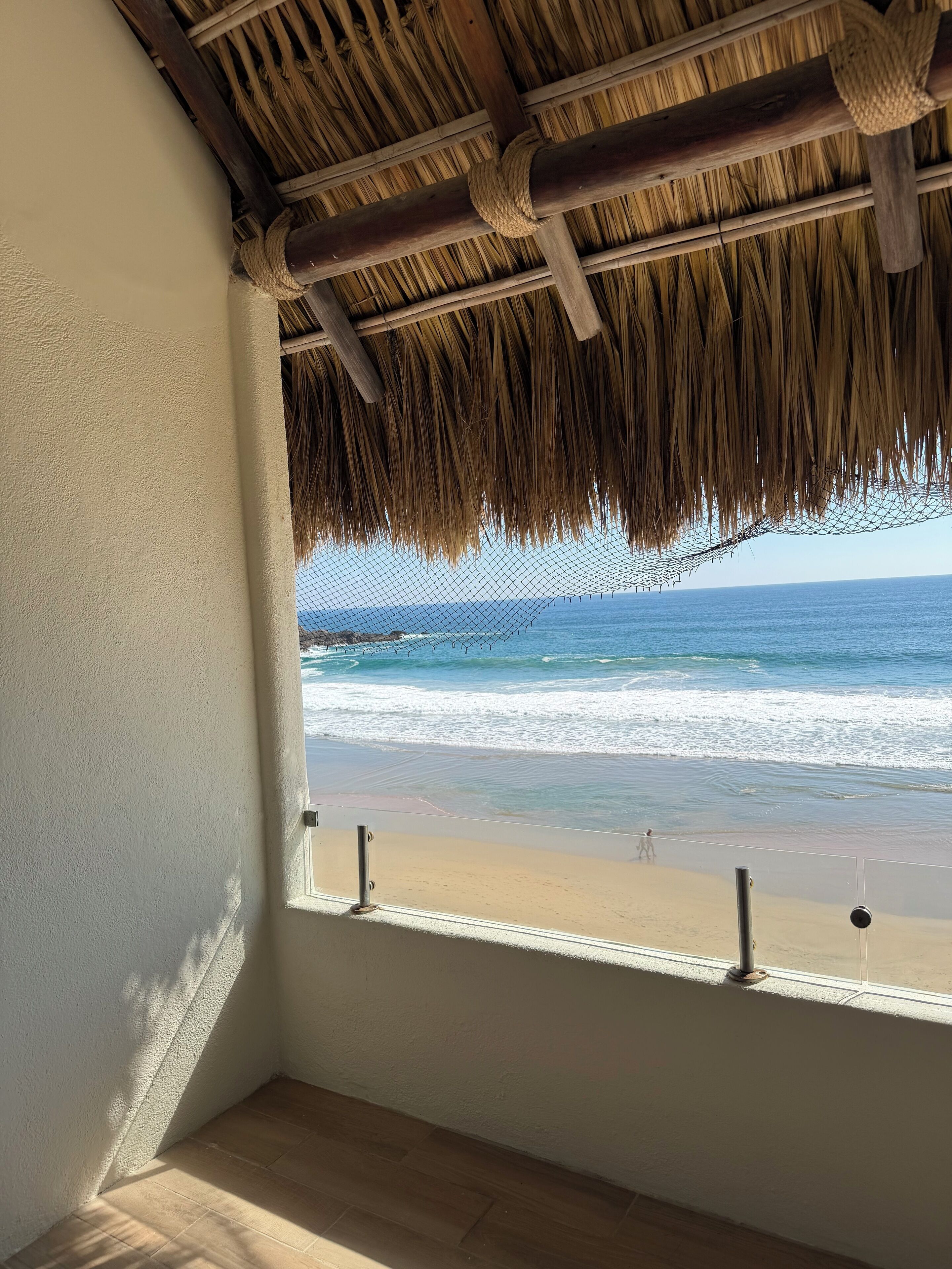 A room with a view of the beach through a window, a thatched ceiling, and a wooden floor.