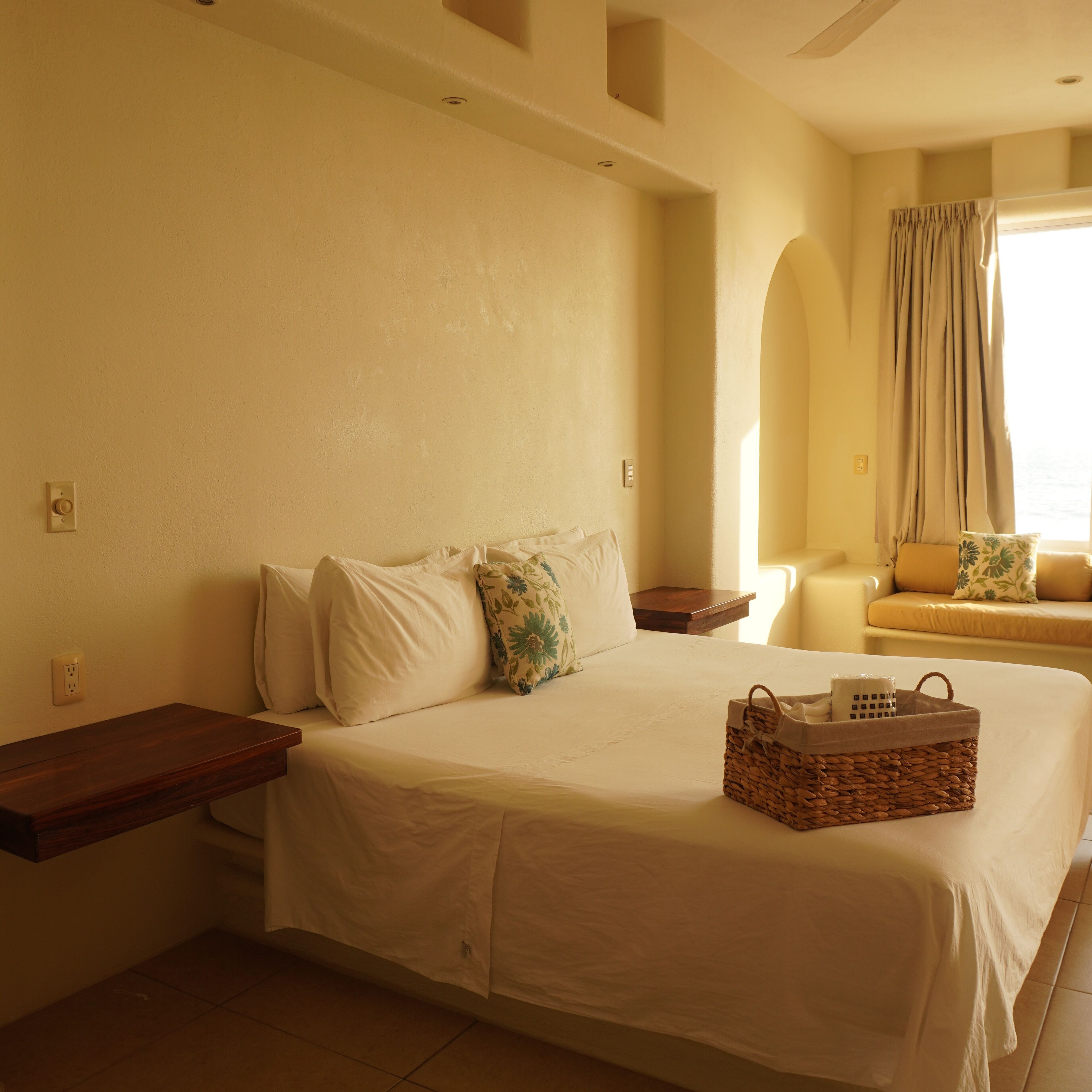 A hotel room with a large bed, a bedside table, a small wooden nightstand, a wicker basket, and a window with curtains.