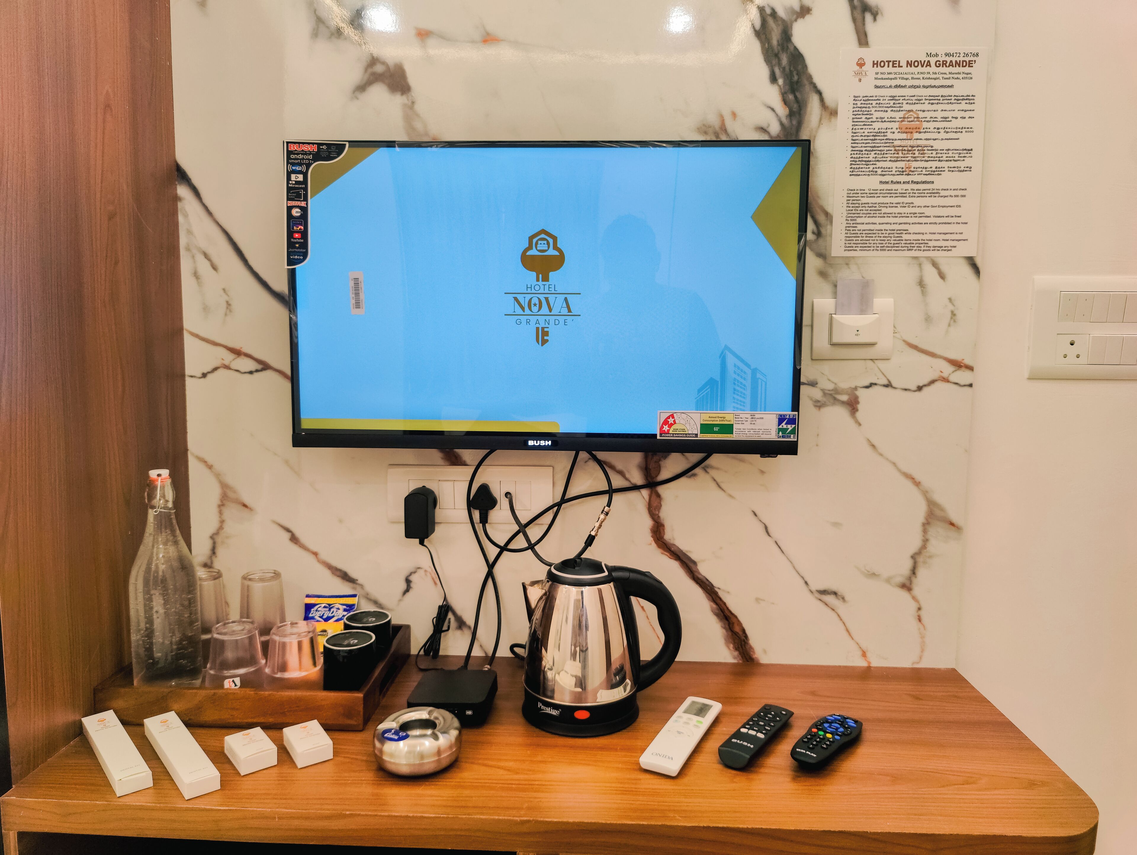 Habitación de hotel con un televisor de pantalla plana mostrando el nombre del hotel, una mesa de madera con varios objetos, un hervidor de agua y controles remotos.