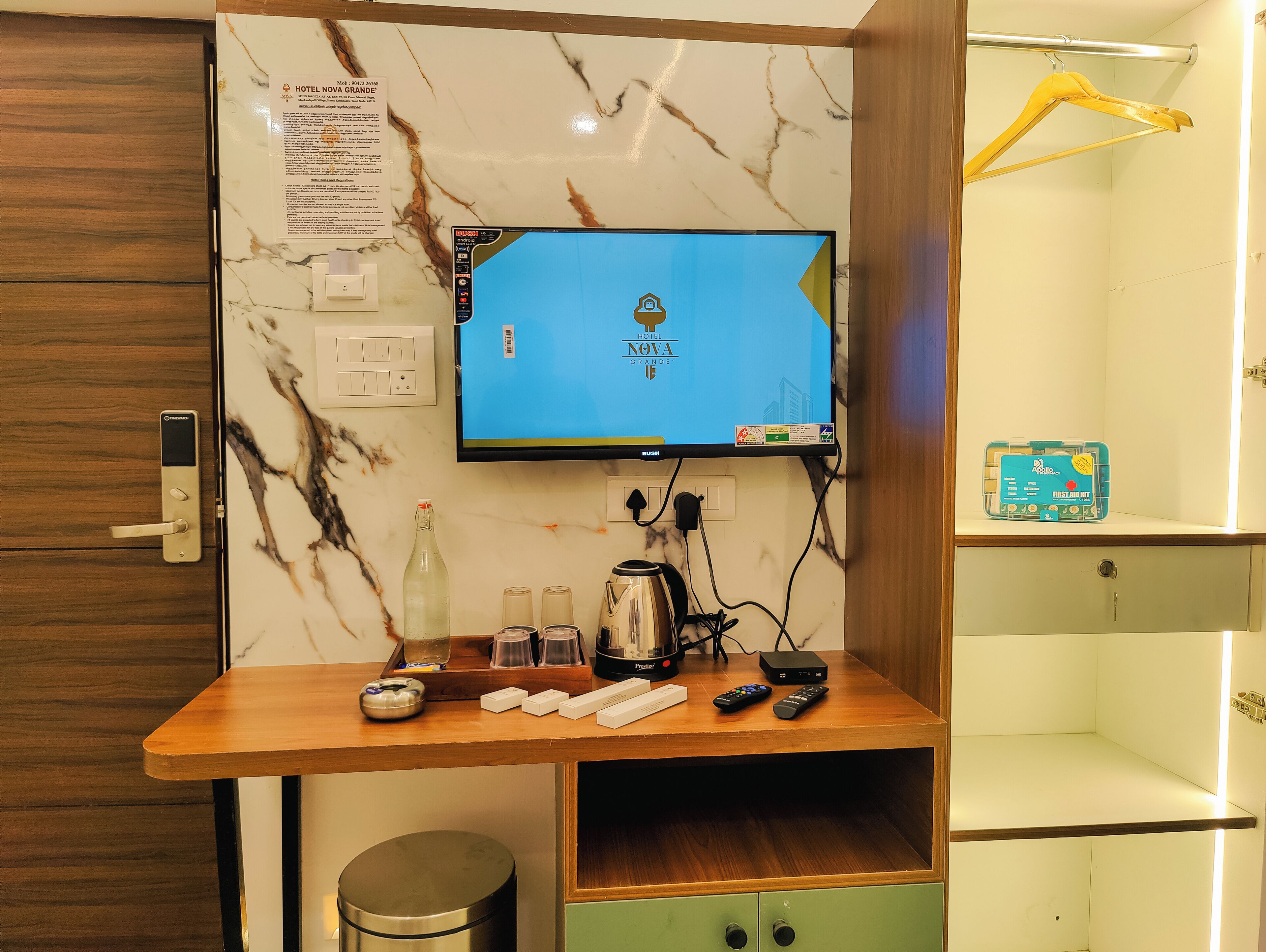 Une chambre d’hôtel avec un mur en marbre, un téléviseur fixé au mur affichant le nom de l’hôtel, un bureau en bois avec une bouteille, des verres, une télécommande et une trousse de premiers secours.