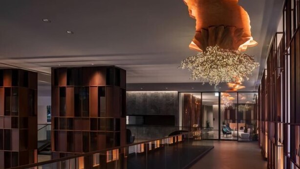 Un hall d’hôtel moderne avec une rambarde en verre, un grand lustre et un espace salon avec des fauteuils.