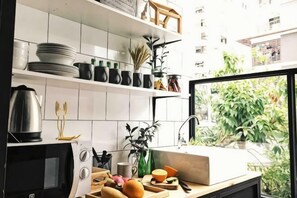 A kitchen with white tiles, a black oven, and a window with a view of greenery. - The Ombra Small House No.4 (Chiang Mai)