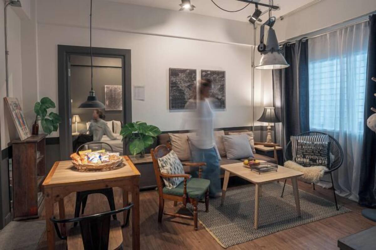 A modern living room with a wooden dining table, a sofa, and a coffee table. There are two paintings on the wall, a potted plant, and a chair with a cushion.