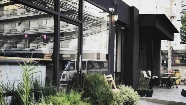 Un bâtiment moderne doté de grandes fenêtres en verre, de plantes en pot et d’un espace extérieur aménagé pour s’asseoir.