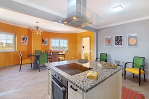 A modern kitchen with a central island, a dining area with chairs, and a view through a window. - Villa 'Picota' with Sea View, Wi-Fi and Air Conditioning (Tavira)