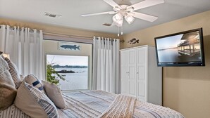 Ein Schlafzimmer mit einem großen Bett, einem Deckenventilator, einem Fernseher und Blick auf ein Gewässer durch ein Fenster.