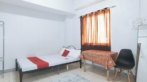 A small, single-bed room with a bed, a table with a patterned tablecloth, a chair, a window with curtains, and a white wall with a decorative piece. - MCP Transient near NAIA (Pasay)