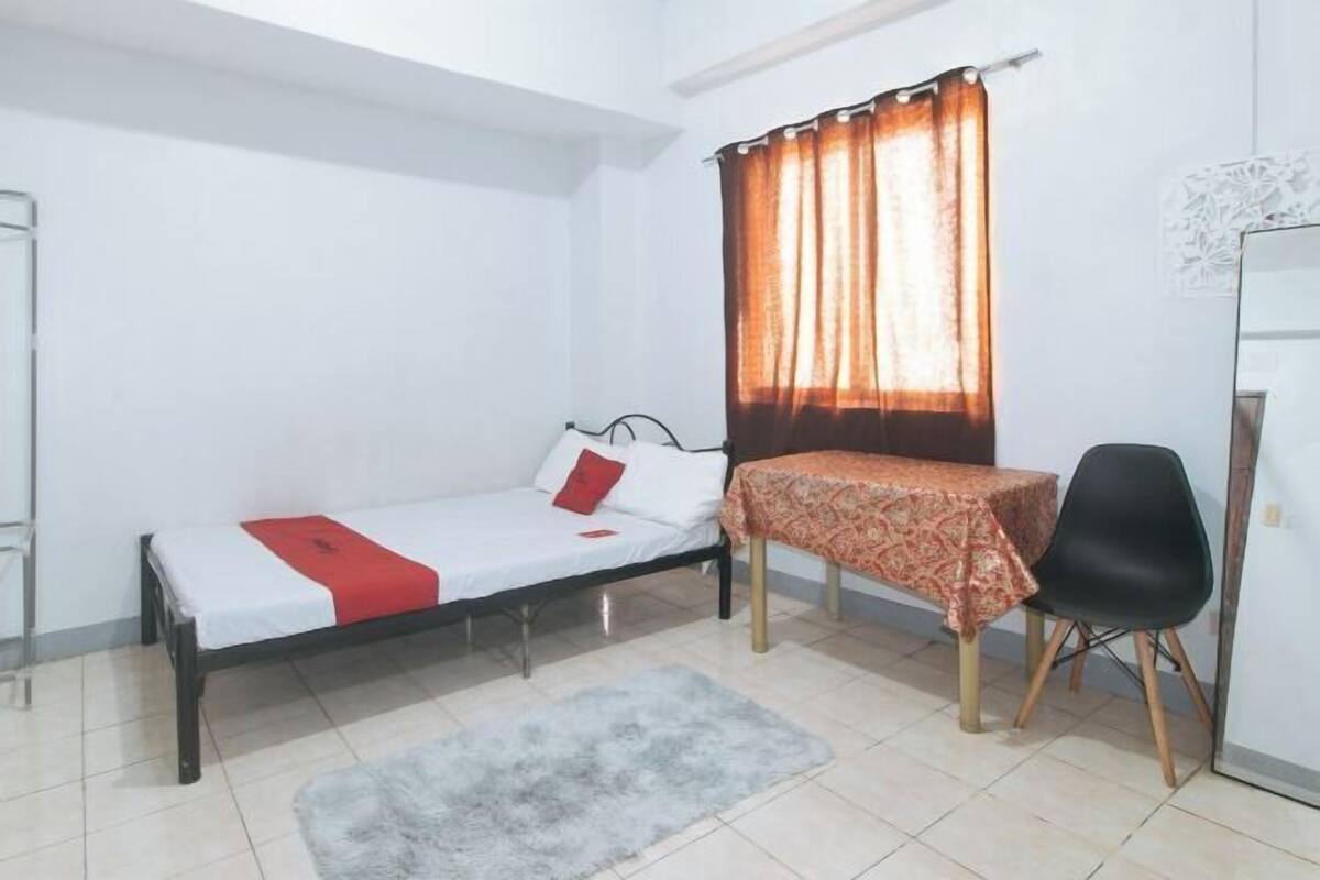 A small, single-bed room with a bed, a table with a patterned tablecloth, a chair, a window with curtains, and a white wall with a decorative piece.