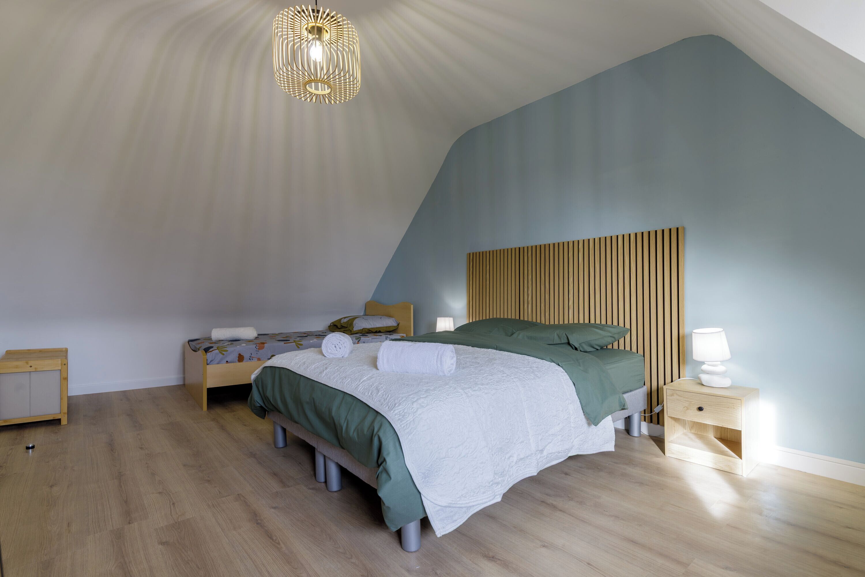 Un dormitorio con techo inclinado, una cama con ropa de cama verde, una mesita de noche con lámpara y un cabecero de madera.