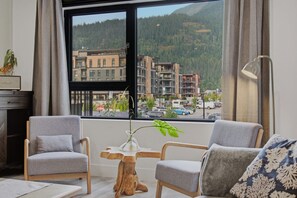 A room with two grey armchairs, a small wooden table, a vase with a plant, and a view of a building complex through a large window.