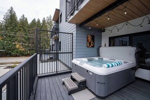 A modern outdoor hot tub on a wooden deck with a railing, surrounded by a building with a grey exterior and a wooden ceiling.