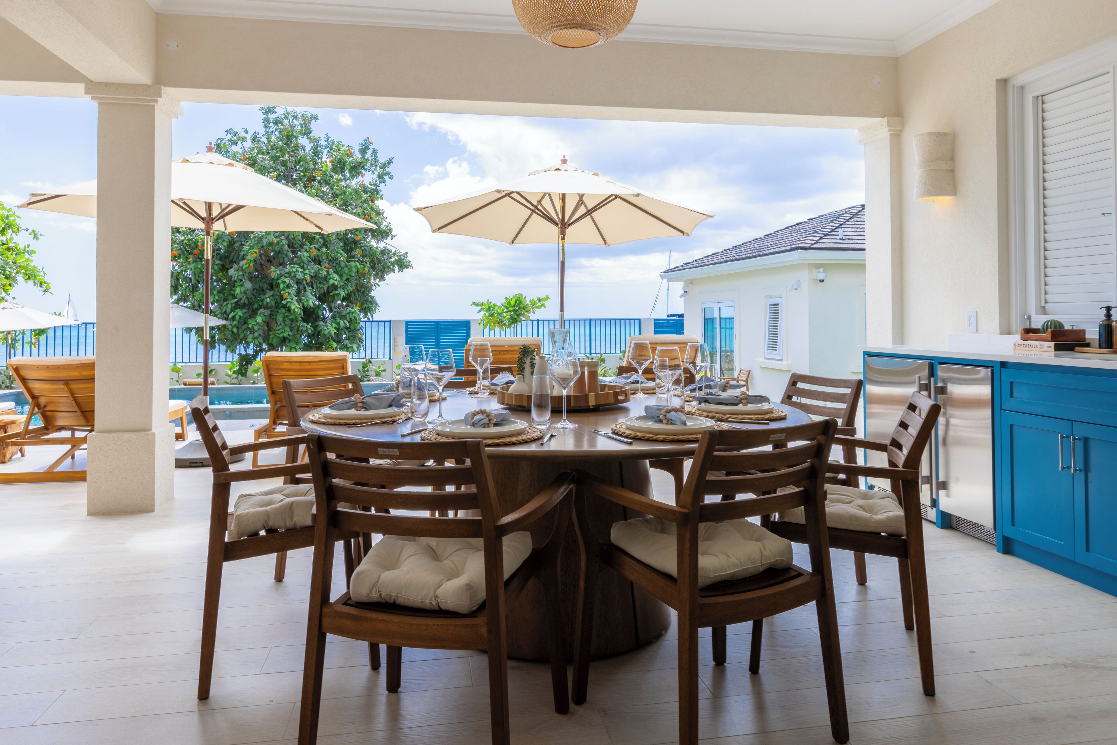 A dining area with a round table set for six, surrounded by wooden chairs with cushions. There are two large umbrellas providing shade.