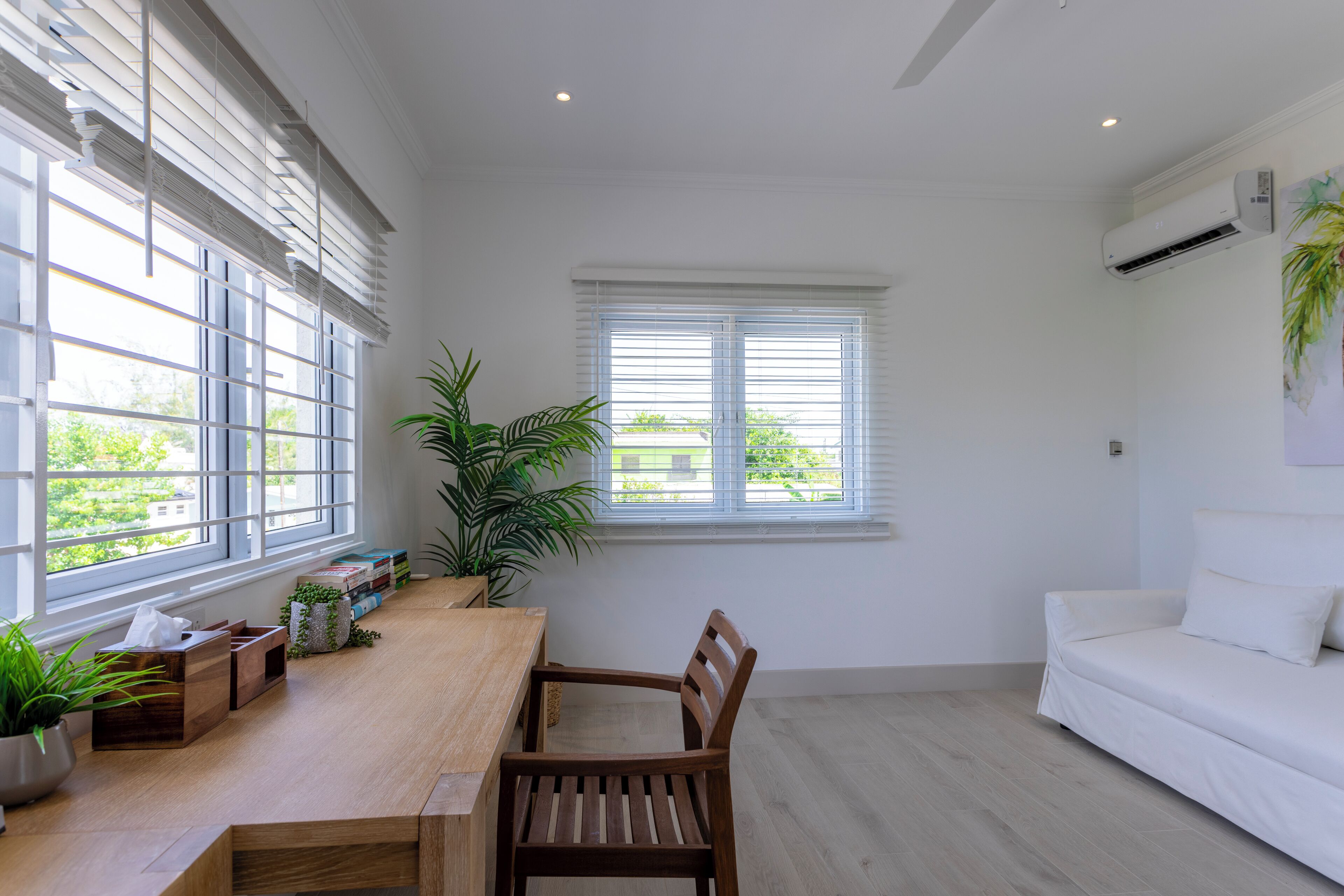 Un salón luminoso y moderno con una mesa de comedor de madera, un sofá blanco y ventanas grandes con persianas.