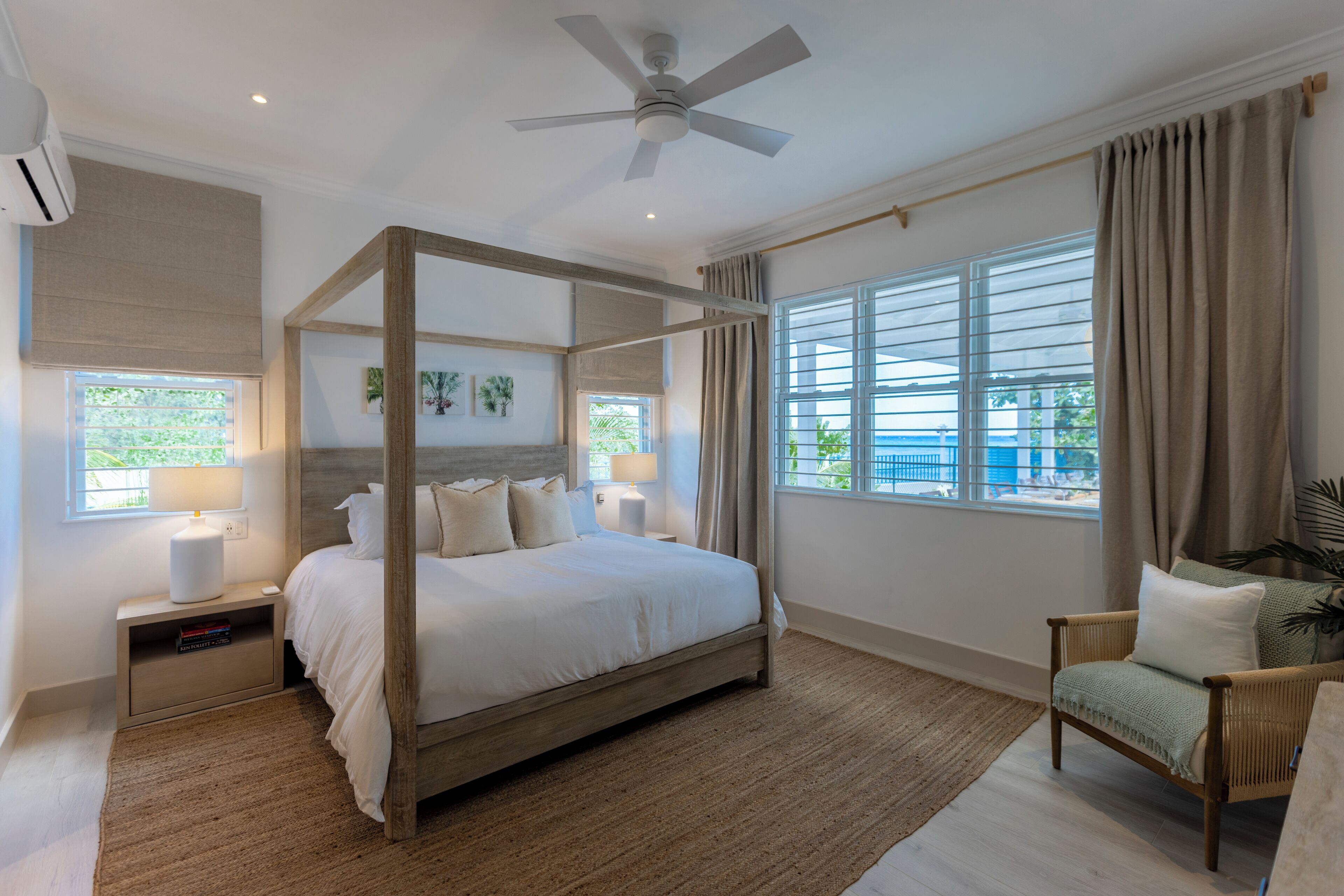 A four-poster bed with a wooden frame, two bedside tables with lamps, a chair, a large window with curtains, and a ceiling fan.