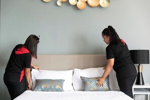 Two individuals arranging a bed with patterned pillows in a hotel room with a neutral wall and a bedside table with a lamp. - Urban Suite@Midhills Genting Highlands (Free WiFi) (Genting Highlands)