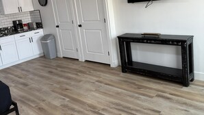 A modern living room with a flat-screen TV, a black console table, and a kitchen area with white cabinets and a coffee maker. - CB&M Westside Suites (Duchesne)