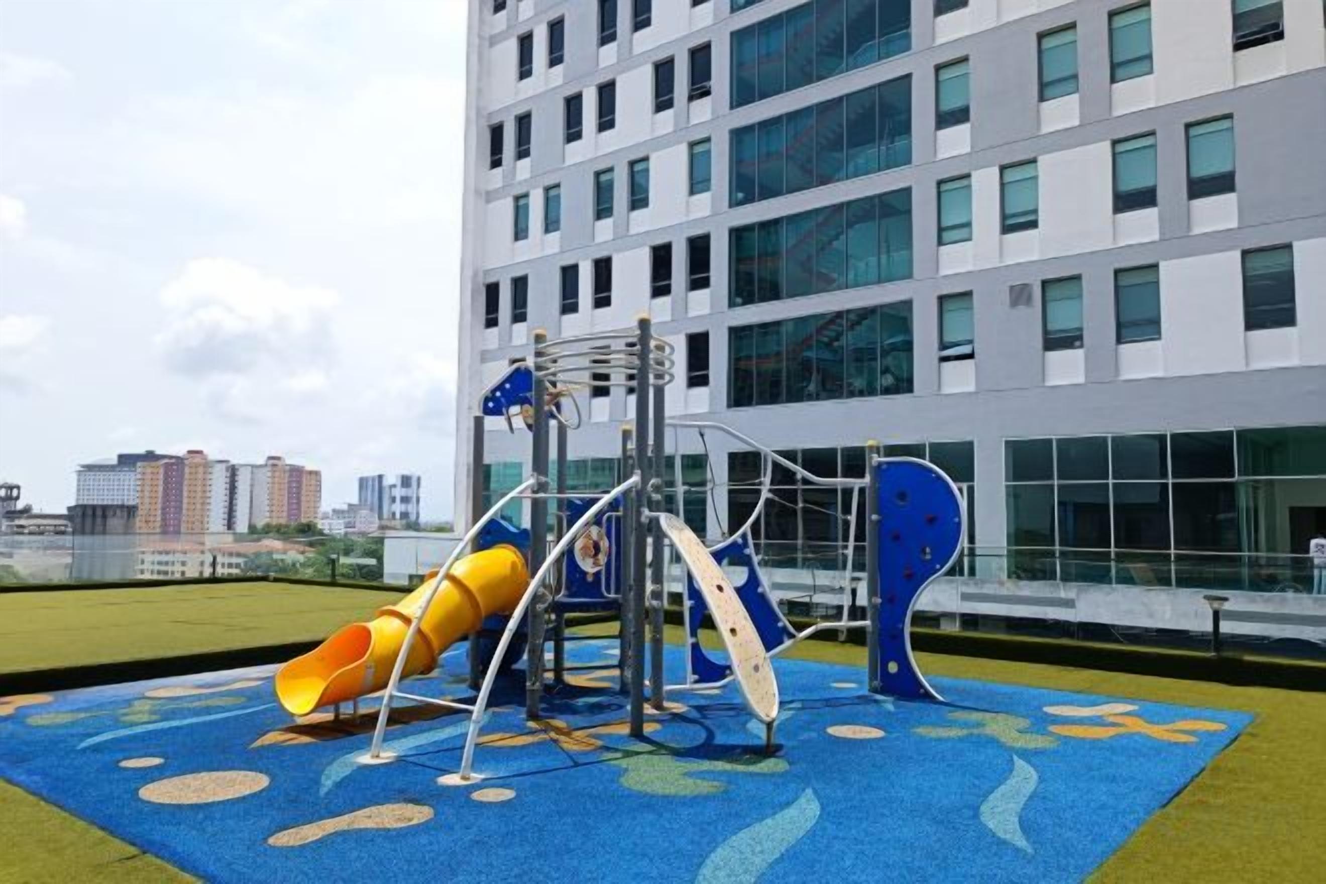 A playground with a slide and climbing structures, located in front of a modern building with large windows.