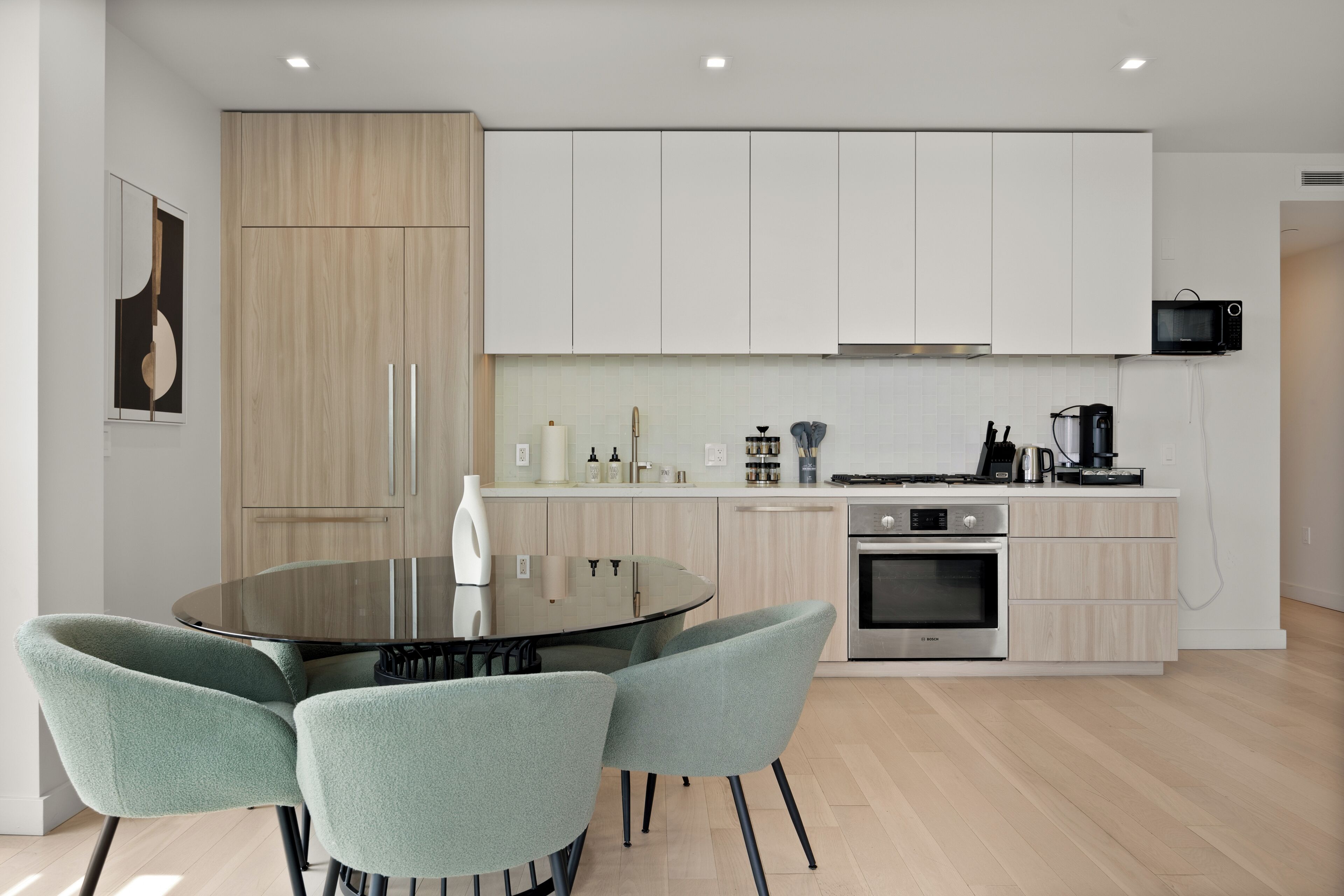 A modern kitchen with white cabinets, a round glass table, and two green chairs.
