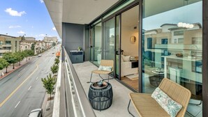 Un balcón con sillas de mimbre y una mesa redonda, con vista a una calle con árboles y edificios.