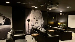 Une pièce avec des canapés en cuir noir, une grande affiche, un écran de projection et une petite table avec une lampe.