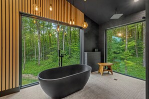Un baño moderno con una bañera negra grande, listones de madera y amplias ventanas con vistas a un bosque.