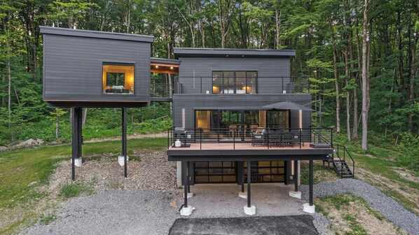 Una casa moderna de dos pisos con balcón y una estructura elevada separada, rodeada por un bosque.