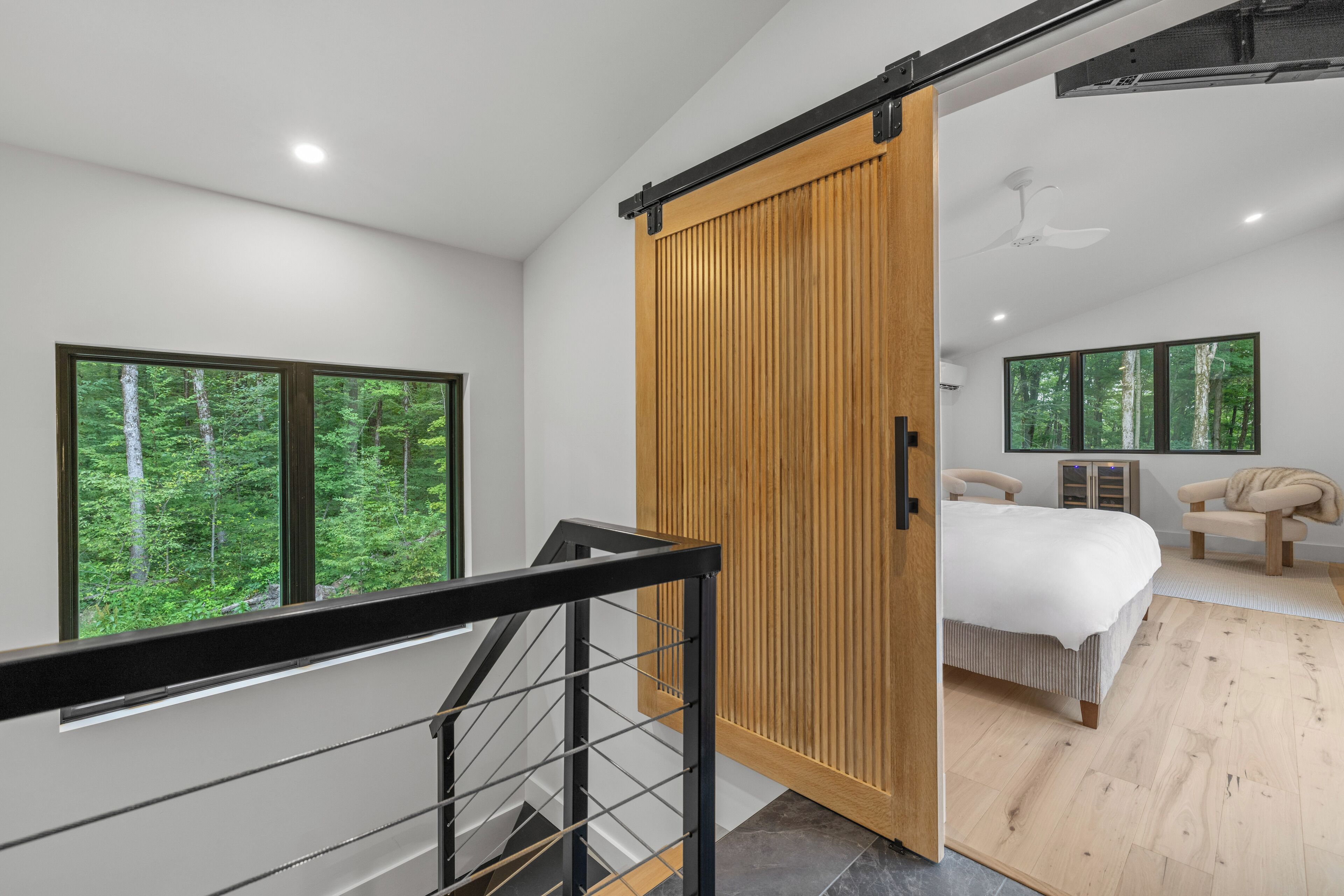 Une chambre moderne dotée d’une grande porte coulissante, d’un lit, d’une chaise et offrant une vue sur une forêt grâce à de grandes fenêtres.