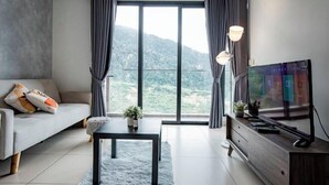 Ein modernes Wohnzimmer mit Sofa, Couchtisch, Fernseher und Blick auf eine gebirgige Landschaft durch große Fenster.