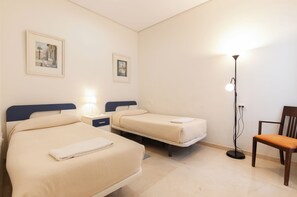 Hotellihuone, jossa on kaksi sänkyä, yöpöytä lampun kanssa, tuoli ja kaksi kehystettyä kuvaa seinällä.