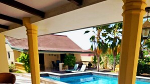 Una mesa de madera con una bandeja que sostiene dos jarrones y un vaso, con vistas a una piscina y tumbonas, y un edificio al fondo.