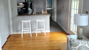 Un vaste espace de vie avec une cuisine à l’arrière-plan, un plancher en bois et une vue sur l’extérieur grâce à une grande fenêtre.