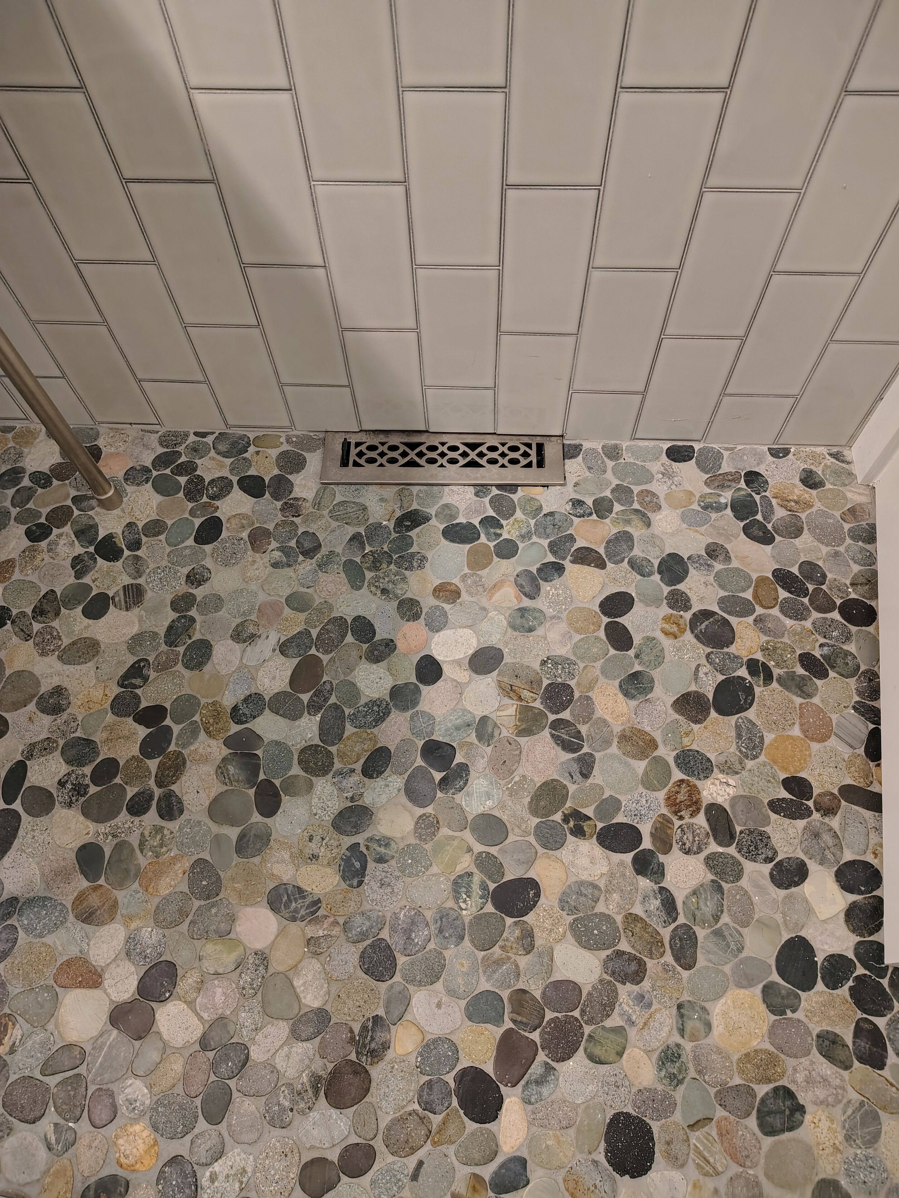 Une salle de bain avec un sol carrelé composé de différents types de carreaux en pierre et un mur également carrelé de blanc, avec un drain au centre.