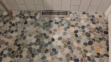 Une salle de bain avec un sol carrelé composé de différents types de carreaux en pierre et un mur également carrelé de blanc, avec un drain au centre.