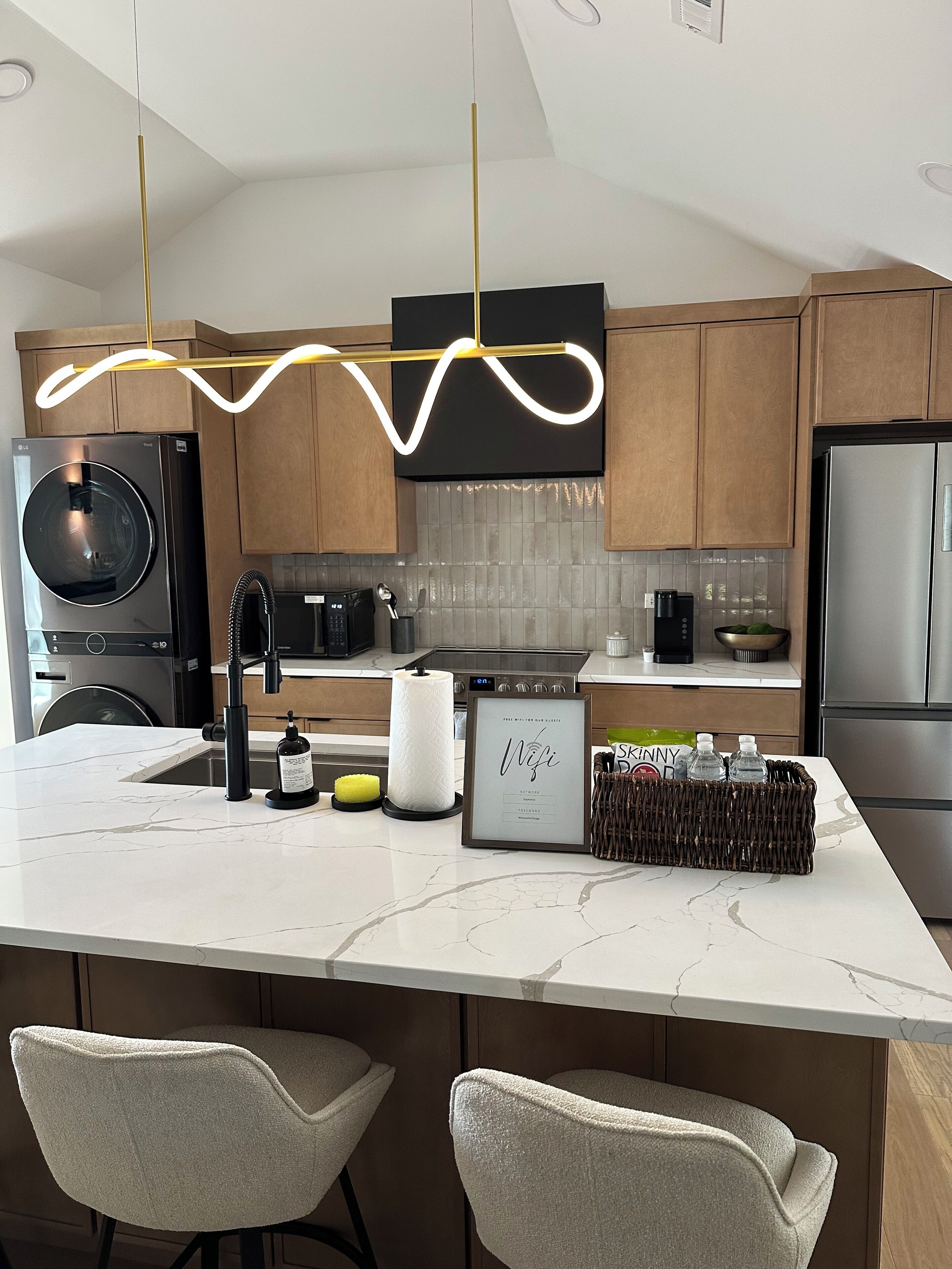 Una cocina moderna con una isla de mármol, armarios de madera, un refrigerador de acero inoxidable y una lámpara colgante.
