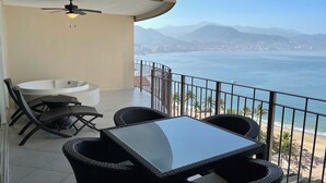 Ein Balkon mit Glastisch und Stühlen, einem Deckenventilator und Blick auf das Meer und die Berge.