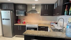Eine moderne Küche mit dunklen Schränken, einem Edelstahl-Kühlschrank, einer Mikrowelle, einem Herd und einer Sp üle. Zwei Barhocker und ein Mülleimer sind ebenfalls vorhanden.