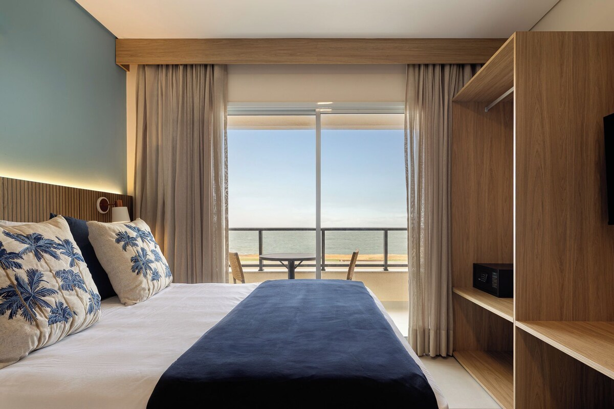 A hotel room with a bed, a TV, a wooden shelf, and a view of the sea.