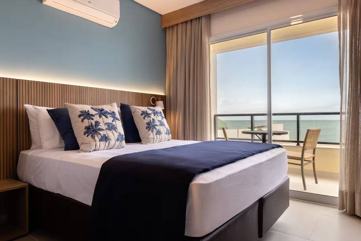 A hotel room with a large bed, a nightstand, a view of the ocean through a balcony, and a ceiling-mounted air conditioning unit.
