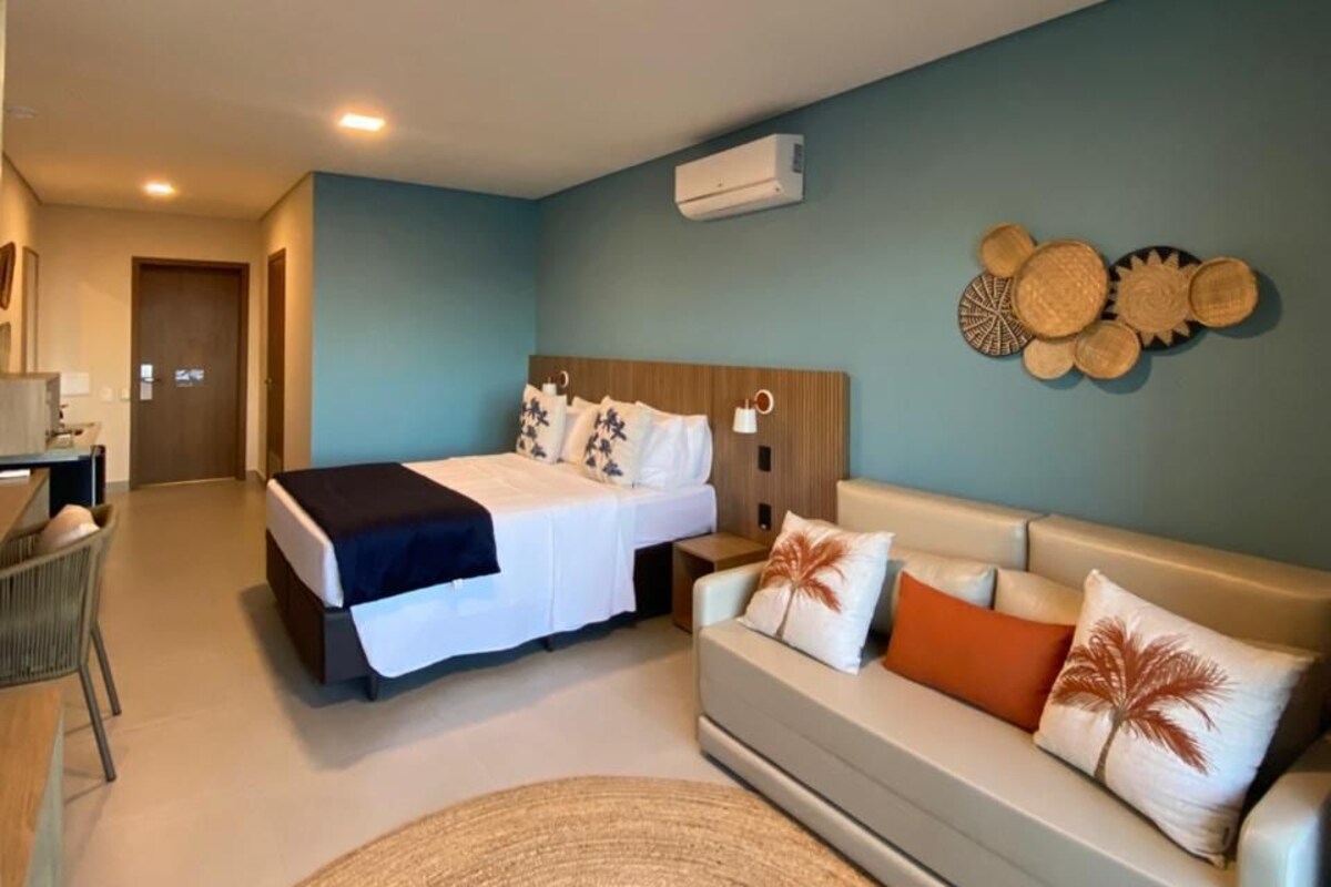 A hotel room with a bed, a sofa, a desk, and a wall-mounted air conditioner.